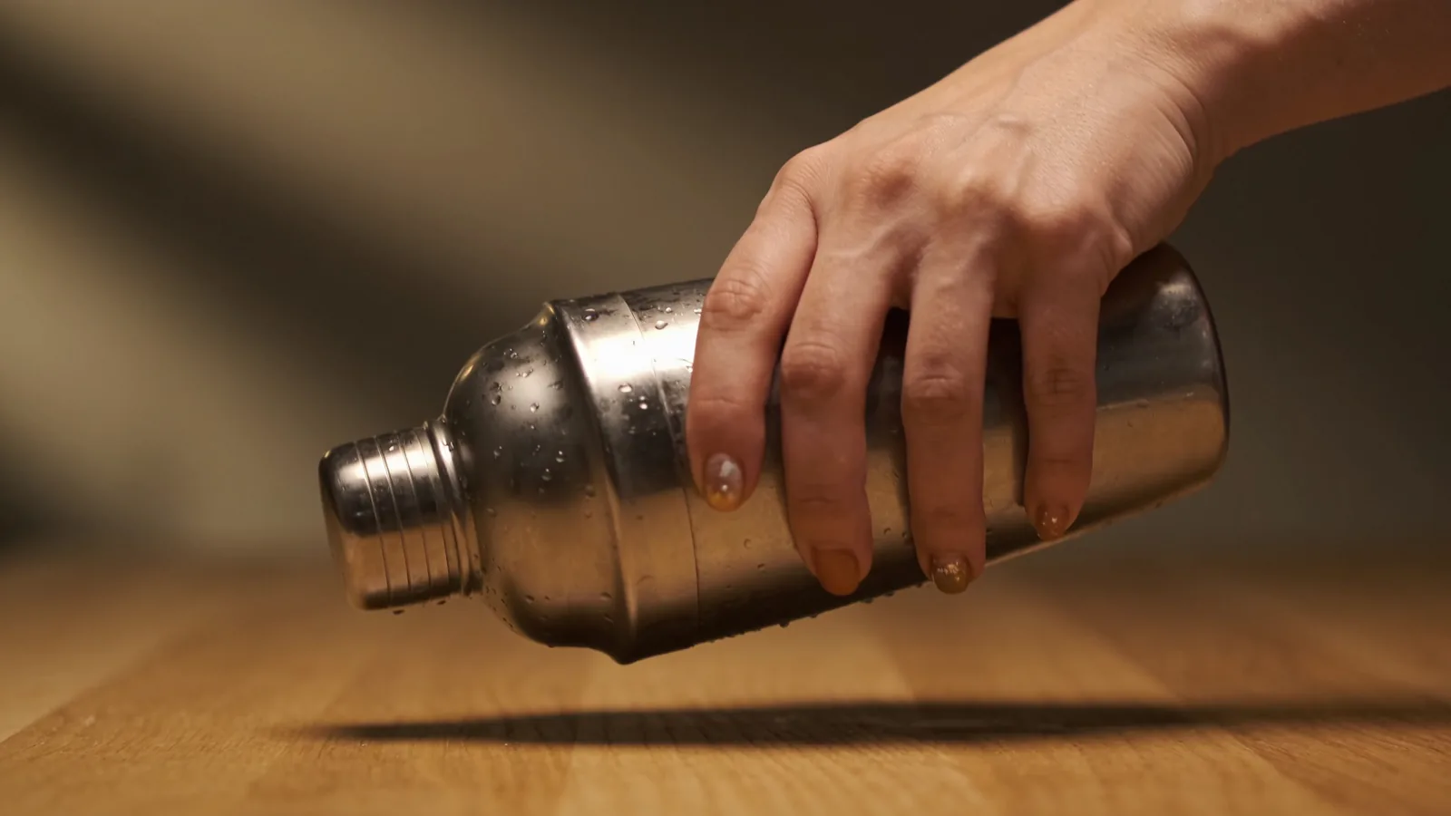 Hand gripping a condensation-covered stainless steel shaker bottle used to mix a protein shake on a wood surface