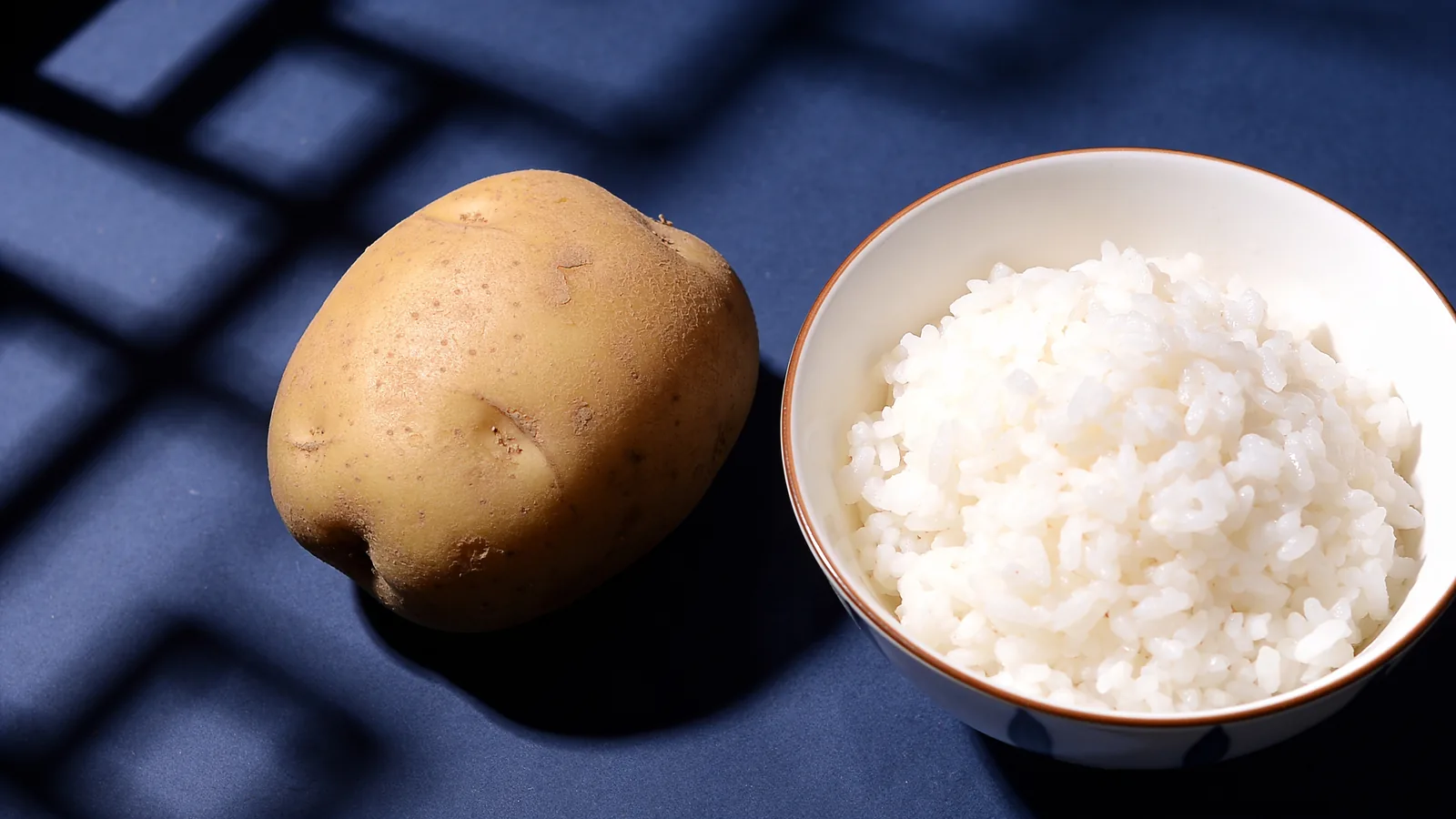 A whole raw potato beside a bowl of cooked white rice on a dark blue surface, source foods for potato vs rice protein