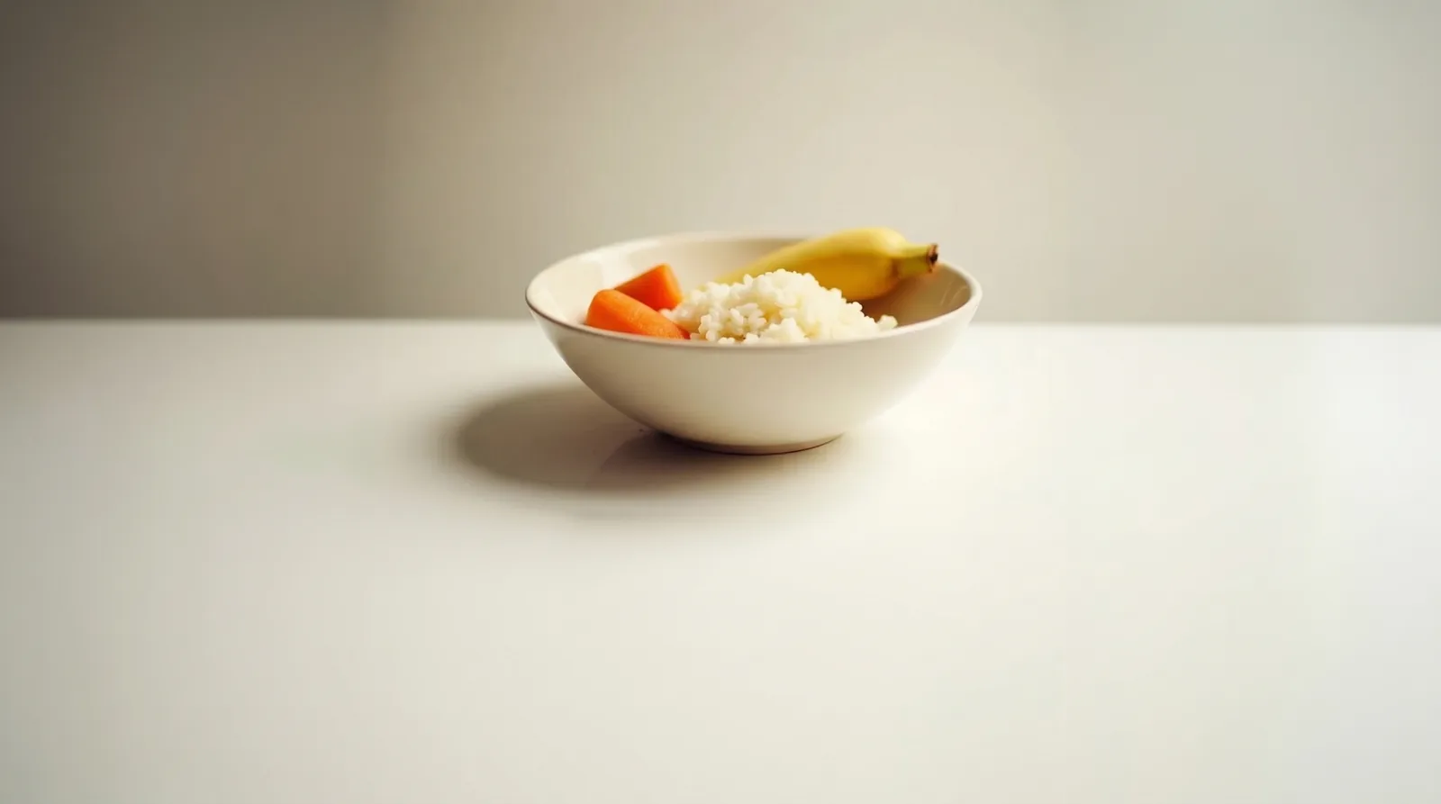 White bowl of cooked rice with carrot pieces and a banana, low-FODMAP foods, on a pale table