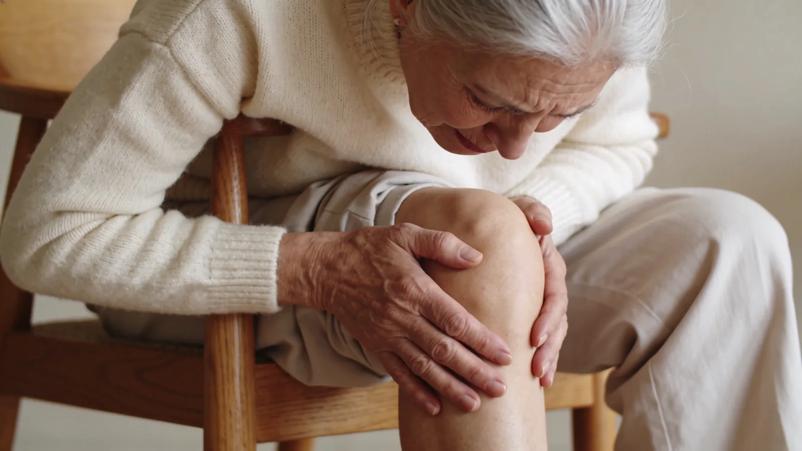 Gray-haired older woman seated on a wooden chair clutching her knee, at an age when bone density declines