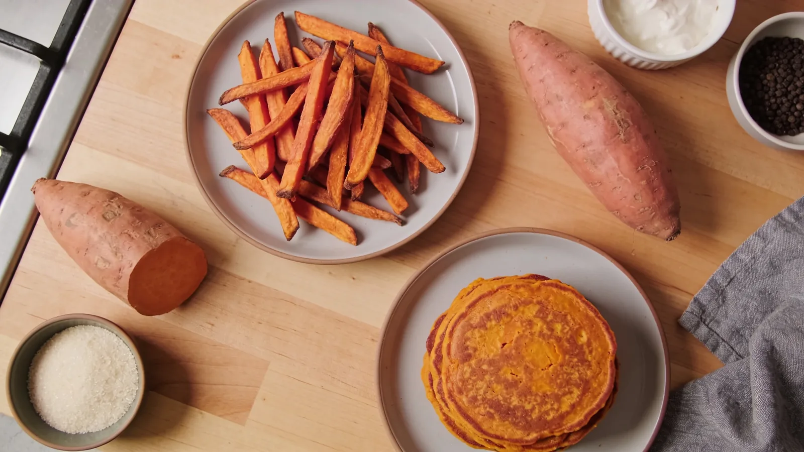 Overhead view of sweet potato fries and a stack of sweet potato pancakes on plates, with raw sweet potatoes, salt, and pepper